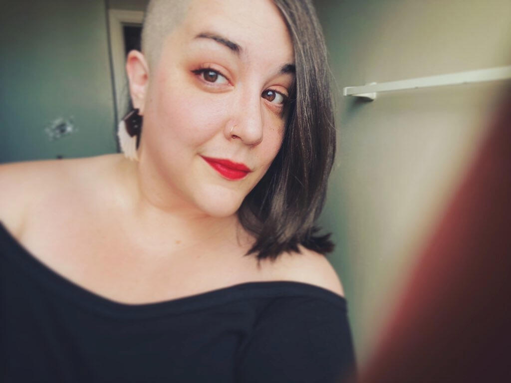 Meredith with a half shave and brown hair, smirks at the camera with red lipstick with a black off the shoulder sweater in a bathroom.